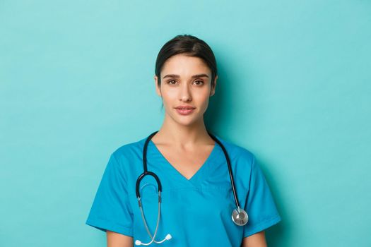 Concept of covid-19 and quarantine concept. Close-up of young confident female doctor with stethoscope, wearing scrubs, standing over blue background
