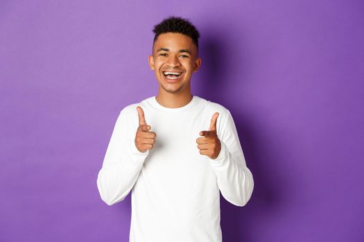Image of cheerful african-american young guy, pointing fingers at camera and smiling, praise good choice, congratulate with something, standing over purple background