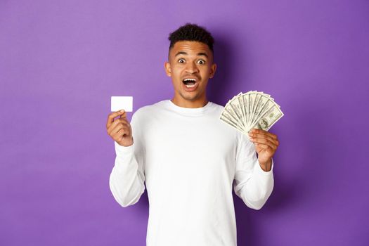 Image of handsome african american guy, looking pleased and smiling, showing credit card and money, standing over purple background