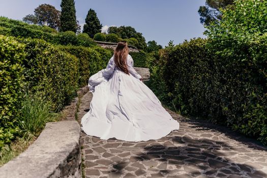 a beautiful woman with long brown hair and long white dress runs along a path along beautiful bushes in the park rear view