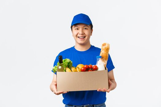 Online shopping, food delivery and shipment concept. Friendly smiling young asian courier in blue uniform handing box with groceries, bring order to customer home, white background
