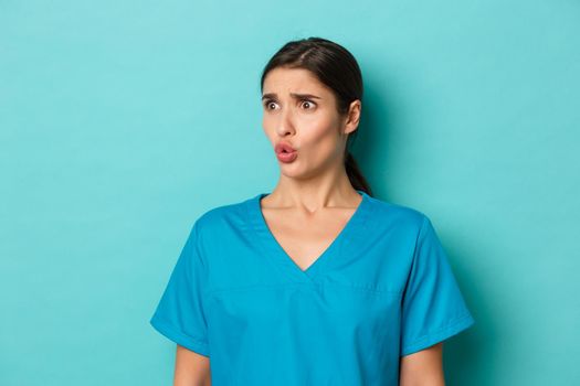 Coronavirus, social distancing and health concept. Close-up of worried brunette female doctor in scrubs, grimacing and looking at something embarrassing, standing over blue background