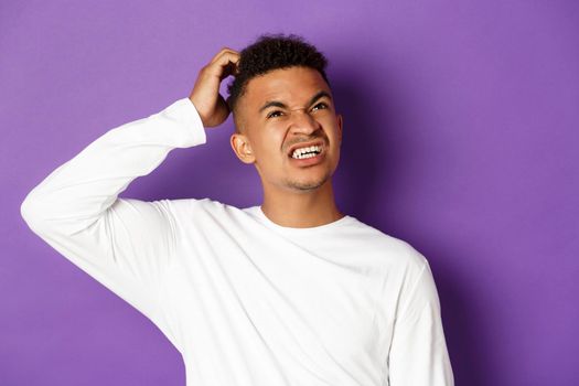 Close-up of troubled african-american man thinking, scratch head and looking up confused, standing in white sweatshirt over purple background