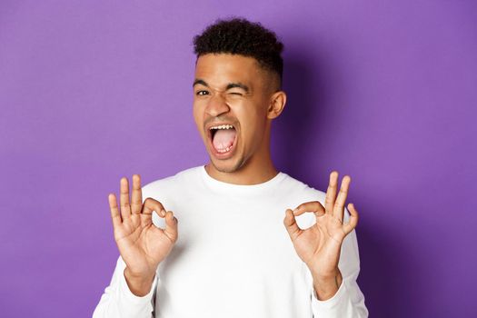 Cheeky african-american man winking, ensure you in something, showing okay signs in approval, standing pleased over purple background