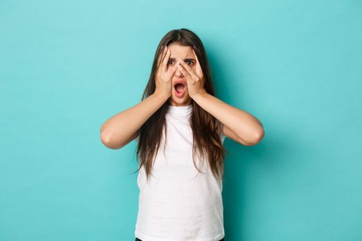 Image of shocked and scared brunette woman in white t-shirt, shut eyes and gasping, peeking through fingers at something embarrassing, standing over blue background
