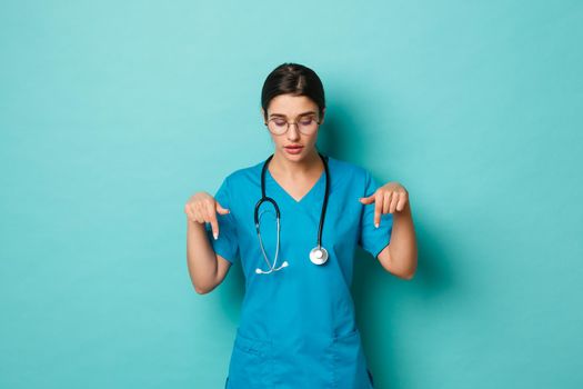 Coronavirus, pandemic and social distancing concept. Image of thoughtful female doctor in scrubs, looking and pointing fingers down curious, standing over blue background
