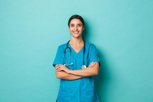 Coronavirus, pandemic and social distancing concept. Image of confident female doctor smiling, wearing scrubs with stethoscope, holding hands crossed on chest, posing over blue background