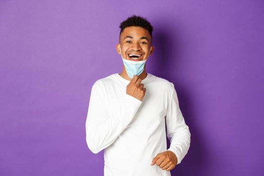 Concept of covid-19, pandemic and social distancing. Happy african-american man taking-off medical mask and breathing freely, standing over purple background