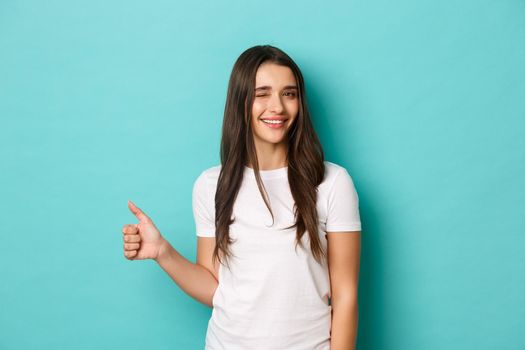 Attractive sassy girl with long dark hair, wearing white basic t-shirt, winking and showing thumb-up, recommend product or company, approve something good, standing over blue background