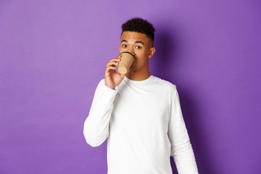 Image of handsome african-american male model, standing over purple background and drinking takeaway coffee