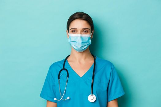 Concept of covid-19 and quarantine concept. Close-up of young female doctor in scrubs and medical mask, looking at camera, standing over blue background