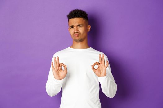 Satisfied handsome african-american man, showing okay signs and looking impressed, praise something good, standing over purple background