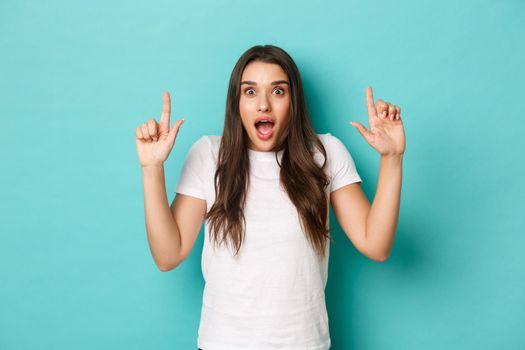 Portrait of surprised and amazed caucasian woman in white t-shirt, gasping and pointing fingers up at something awesome, standing over blue background