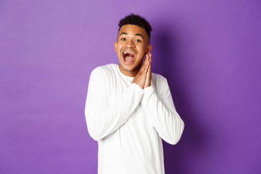 Image of excited and hopeful african-american queer guy, looking with anticipation and thrill at something, clap hands and smiling happy, standing over purple background