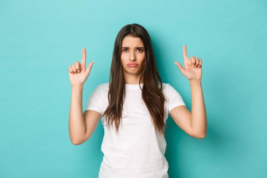 Portrait of sad and gloomy cute girl in white t-shirt, grimacing upset and pointing fingers down, showing something bad, standing over blue background