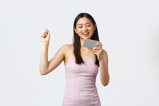 Luxury women, party and holidays concept. Happy satisfied pretty woman in evening dress, do champion dance as winning in mobile game, holding smartphone and fist pump excited, white background