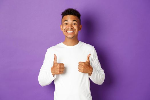 Handsome cheerful man in white sweatshirt, showing thumbs-up in approval and smiling, like and praise something good, standing over purple background
