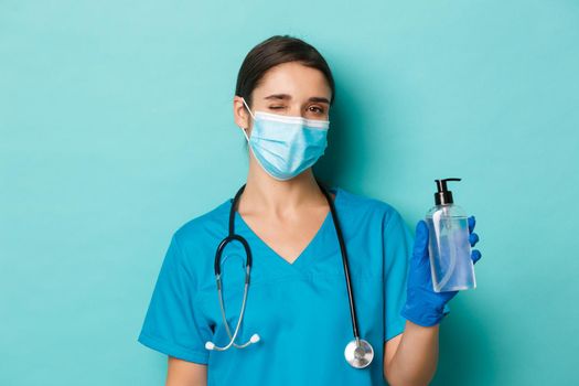 Concept of covid-19 and quarantine concept. Close-up of beautiful female doctor in medical mask, gloves and scrubs, winking and showing hand sanitizer, standing over blue background