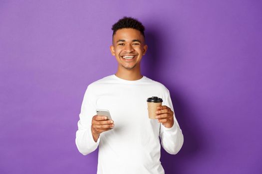 Portrait of handsome african-american guy smiling pleased, drinking coffee and surfing the net in mobile phone, standing over purple background