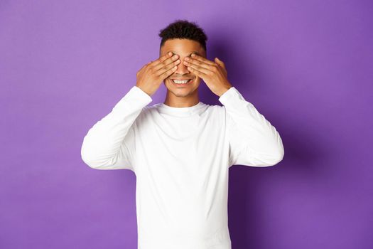 Image of handsome african-american 20s guy, cover eyes with hands and smiling, waiting for surprise with excited face, standing over purple background