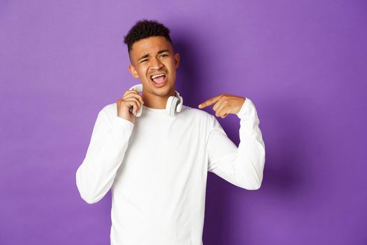 Image of sassy african-american guy, pointing at headphones and praising awesome song, standing over purple background