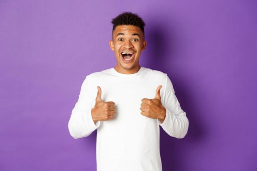 Excited african-american man showing thumbs-up, smiling amazed, like and praise great promo, standing over purple background