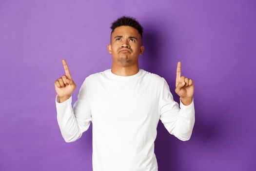 Disappointed and sad african-american young guy in white sweatshirt, looking and pointing fingers up, showing something bad, sulking upset over purple background