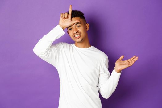 Image of arrogant and confident african-american man, mocking lost team and showing loser sign on forehead, standing over purple background