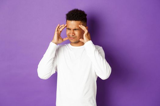 Image of handsome african-american man having headache, grimacing and touching head, suffering migraine, standing over purple background