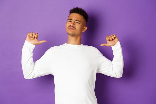 Image of confident and handsome african-american man, pointing at himself with proud face, standing self-assured over purple background