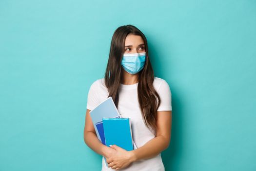 Concept of coronavirus, health and social distancing. Portrait of female student attend classes in medical mask, looking right at copy space, holding notebooks, standing over blue background