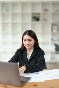 Portrait of a beautiful Asian businesswoman in a formal suit sits at her office desk with using notebook and laptop computer in office