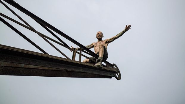 Shirtless muscular man sits on an old rustic crane shouts