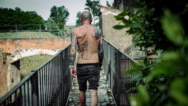 An exhausted man with body covered with tattoos walks on a narrow bridge