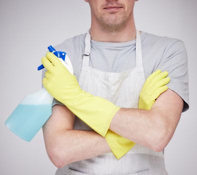 What needs cleaning today. an unrecognizable man holding a spray bottle with his arms crossed against a grey background.