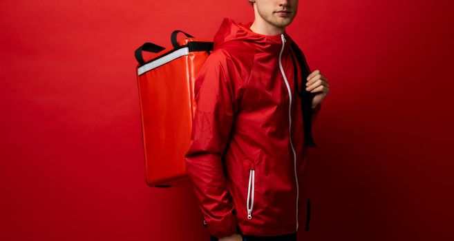 Delivery employee man in red uniform and thermal food bag backpack work courier service on red background studio.