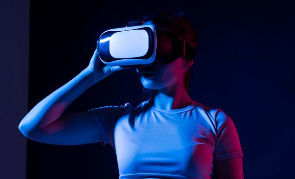 Young woman using VR goggles in colorful neon lights and having fun playing a games with a friends. Wearable virtual augmented reality.