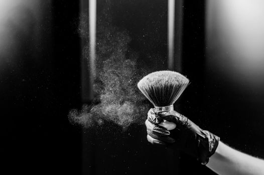 A woman shakes a large powder brush with clouds of dust on a black background. Treatment after instant tanning