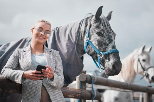 Fresh air and back rides. Portrait of an attractive woman using her cellphone while posing with a horse in an enclosed pasture on a farm.