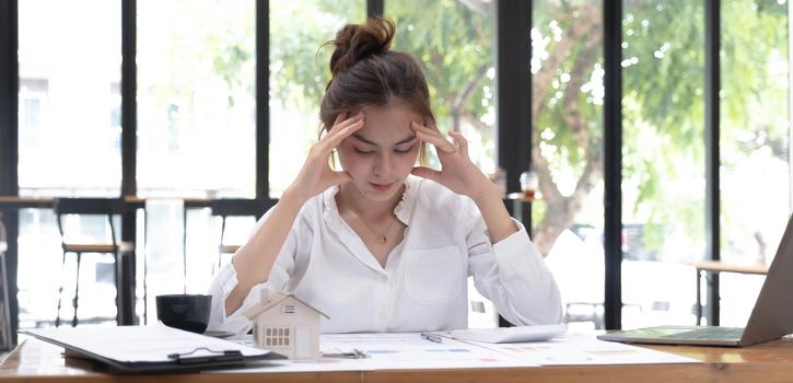Photo of a tired woman keeps a hand on her head at the wooden working desk after finishing work hard.