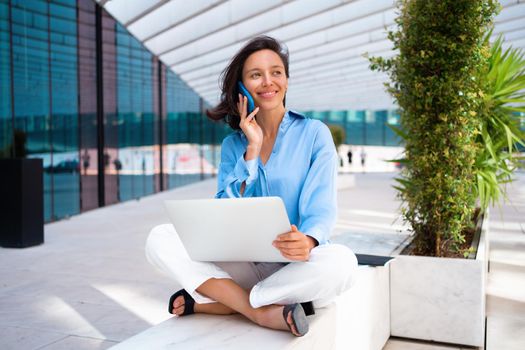 Self employed woman working with her phone and laptop sitting outdoor near corporative office building. Pretty business woman freelancer do remote work sitting lotus position