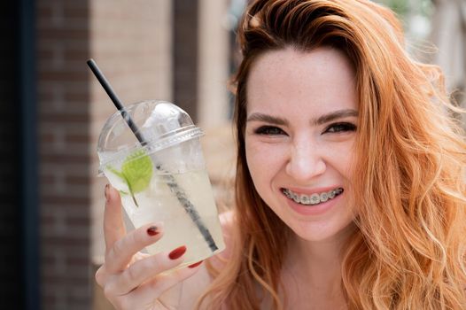 Young beautiful red-haired woman with braces drinks cooling lemonade outdoors in summer. Portrait of a smiling girl with freckles.