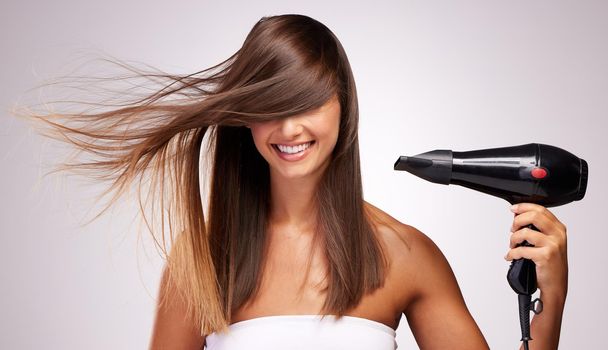 Lets get this hair dried. Studio shot of an attractive young woman blowdrying her hair against a grey background.