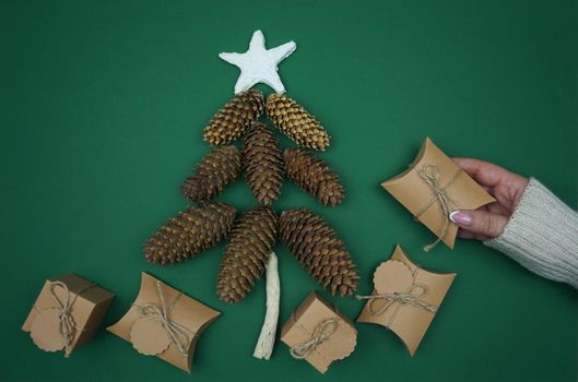 On a green background, a woman's hand laid out gifts in craft paper and made a Christmas tree out of fir cones