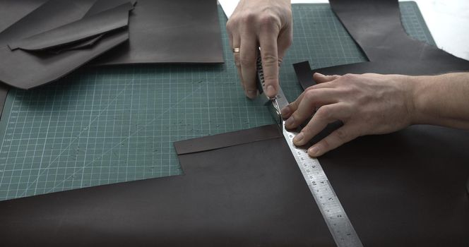 Close up of leather craftsman working with natural leather by knife and ruler by cutting a leather to a peaces. Handmade master at work in local workshop. Leather wallet stitch.