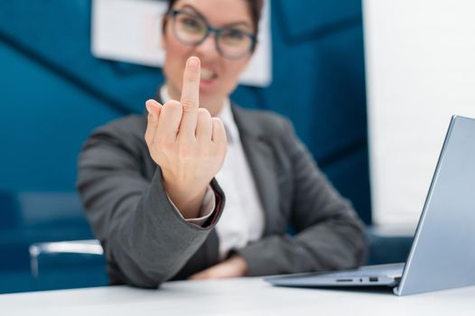 Angry business woman in a suit shows a middle finger while sitting at a desk. Annoyed female office worker showing a fuck you gesture. Professional burnout.
