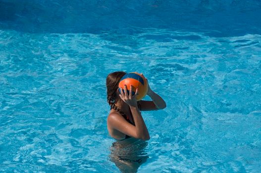 Young tanned girl holding a ball and playing water volleyball in the pool