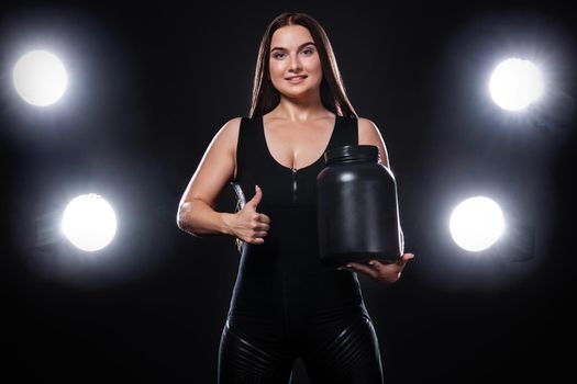 Happy and healthy muscular young fitness sports woman athlete with a jar of sports nutrition - protein, gainer and casein on black background.
