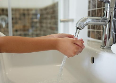 A closeup of a child washing her hands at the sink inside a bathroom with running tap water. A kid practicing hygiene and cleanliness by cleaning her fingers for virus prevention