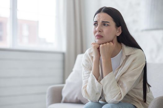 Depressed woman near window sitting on sofa at home, disappointed and sad brunette grieving divorce